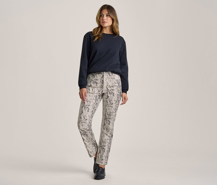Femme debout, portant un haut à manches longues bleu foncé, un pantalon gris clair à motif peau de serpent noir et des chaussures noires.