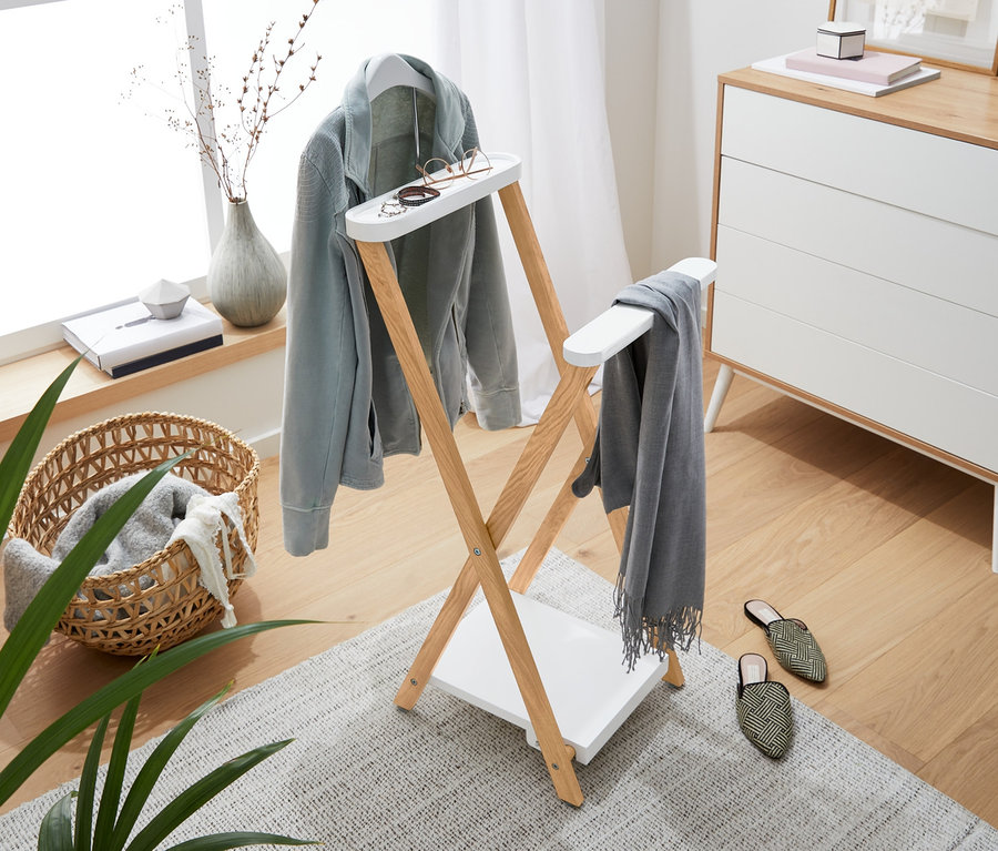 Pièce lumineuse avec un portant en bois sur lequel sont suspendus une veste bleu clair, une écharpe grise et des lunettes. En arrière-plan, une commode, une fenêtre et des décorations.