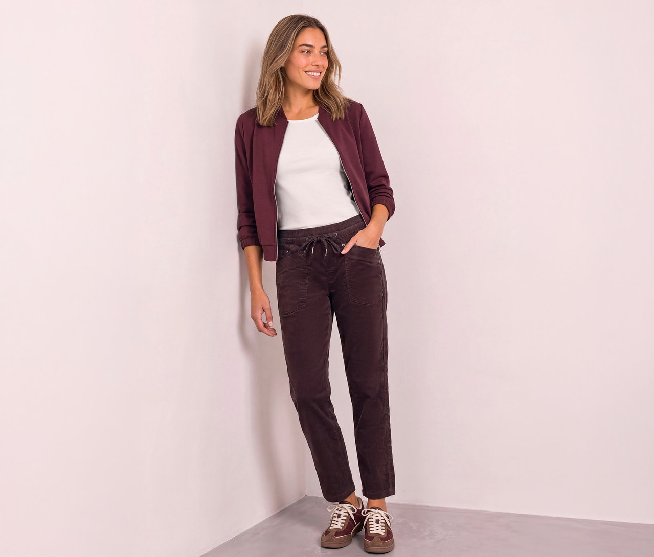Une femme souriante aux cheveux mi-longs, vêtue d'un blouson bomber bordeaux sur un t-shirt blanc, d'un pantalon marron foncé à cordon de serrage et de baskets marron, s'appuie contre un mur blanc, les mains dans les poches.