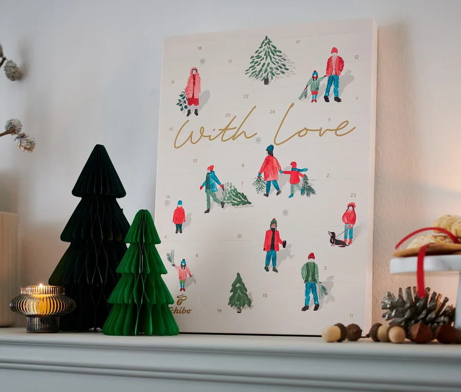 Sur une étagère sont disposés des objets de décoration, dont un Calendrier de l'Avent Tchibo, un vase avec des branches, des sapins en papier et une bougie.