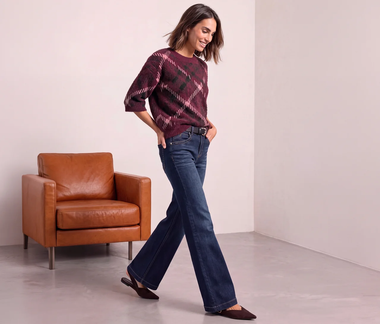 Femme portant un pull en maille Street One à motif losanges bordeaux, rose et vert foncé, un jean évasé bleu foncé et des chaussures plates marron, marchant devant un fauteuil en cuir marron.