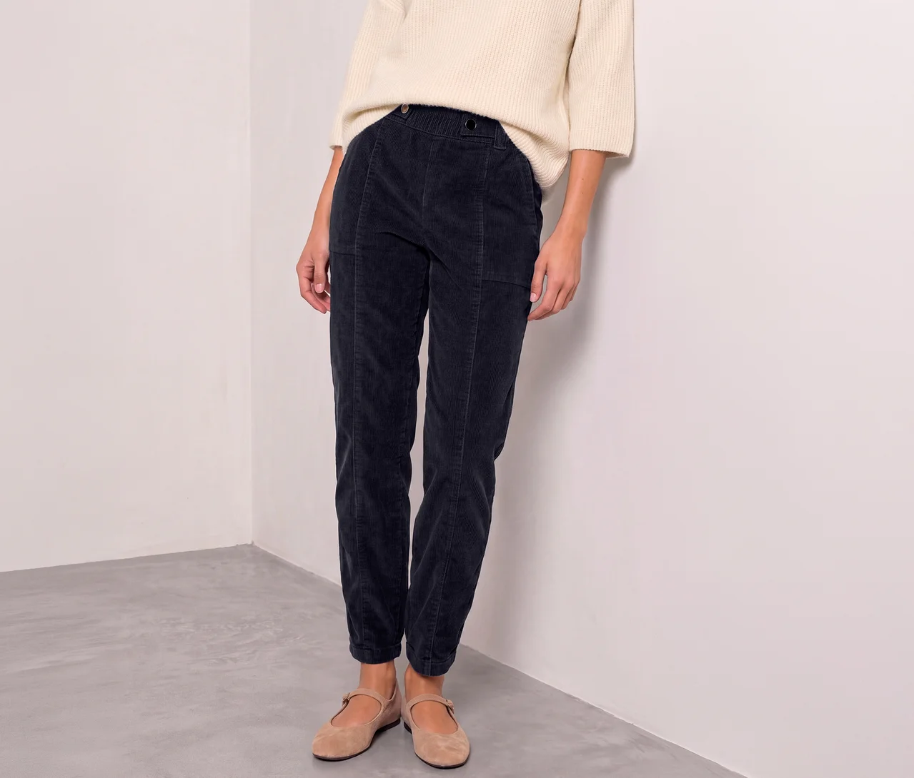 Une personne porte un pull crème, un pantalon en velours côtelé Street One bleu foncé et des ballerines marron clair, adossée à un mur blanc.