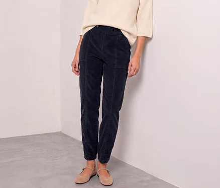 Une personne porte un pull crème, un pantalon en velours côtelé Street One bleu foncé et des ballerines marron clair, adossée à un mur blanc.