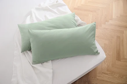 Deux taies d’oreiller en jersey vert sauge sont posées sur un drap blanc.