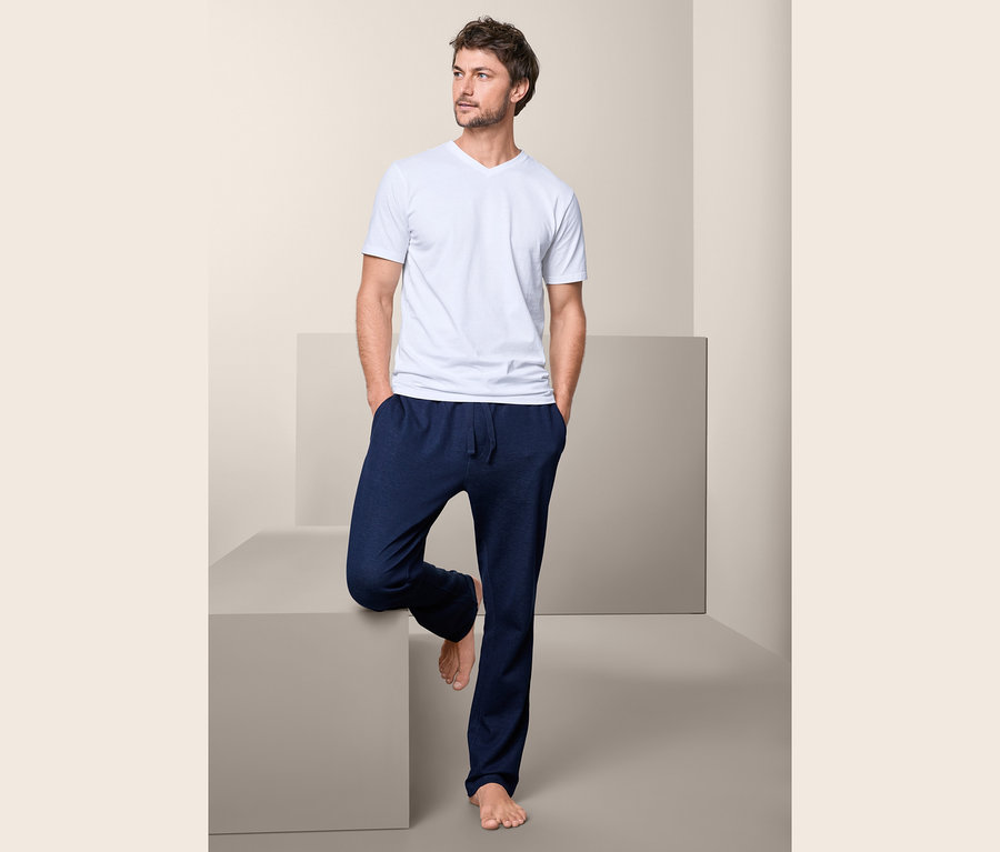 Un homme portant un t-shirt blanc à col en V et un pantalon bleu est appuyé contre un cube gris.