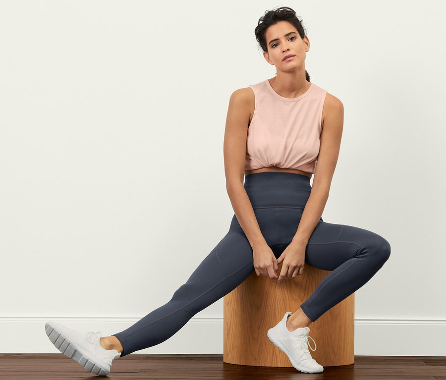 Une femme est assise sur un tabouret en bois et porte un legging de sport « Sculpture », un haut et des baskets.