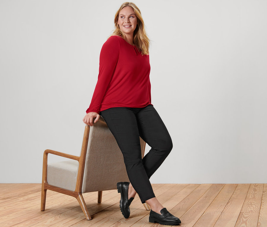 Une femme est assise sur une chaise et porte un chemisier rouge, un pantalon en bengaline noir et des mocassins en cuir.