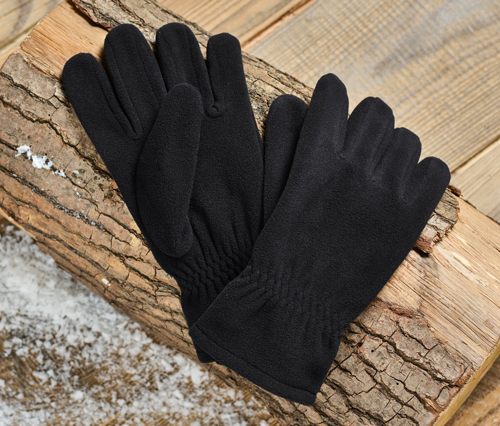 Une paire de gants en micropolaire noirs posés sur une bûche en bois sur un fond en bois.