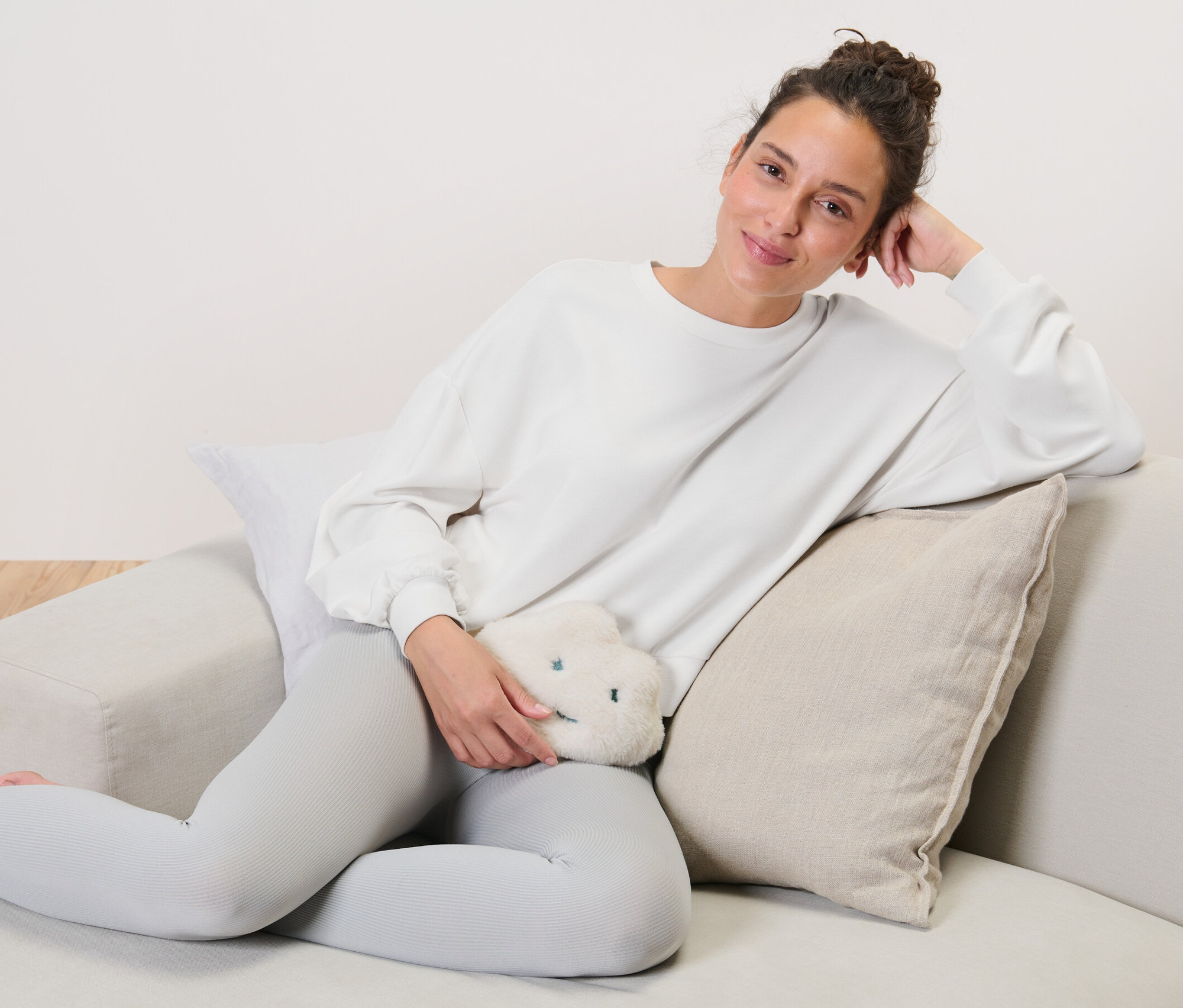 Une femme est assise sur un canapé et tient un coussin chauffant aux noyaux de cerise en forme de nuage.