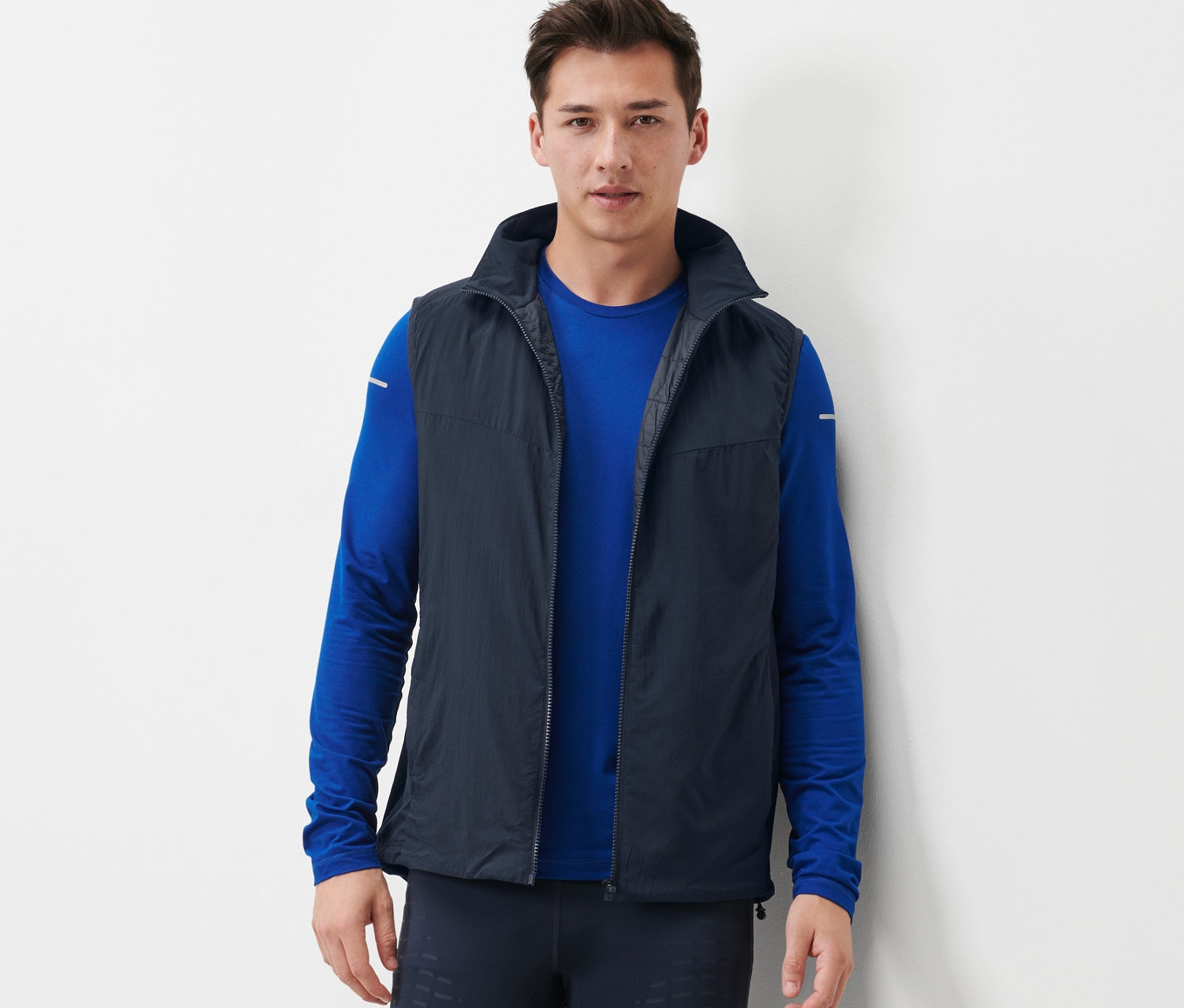 Un homme portant un gilet technique sans manches, un t-shirt bleu à manches longues et des leggings foncés.