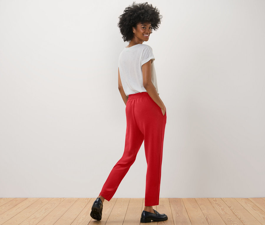 Une femme souriante pose en pantalon de jogging rouge et un t-shirt blanc.