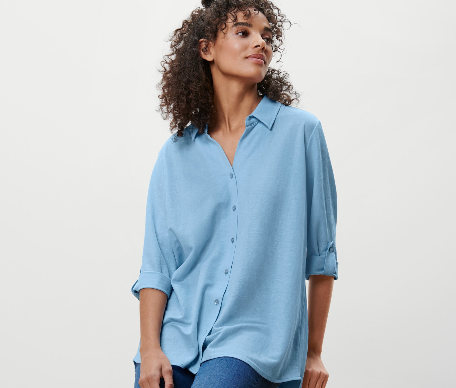 Une femme est assise sur un tabouret et porte un chemisier en jersey piqué bleu clair et un jean.