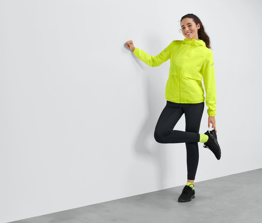 Une femme posant contre un mur, portant une veste de sport coupe-vent jaune.