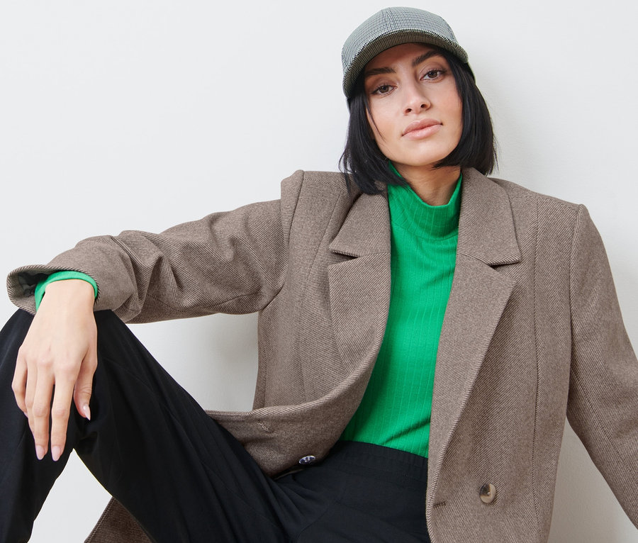 Une femme est assise et porte un blazer, un col roulé vert et une casquette.
