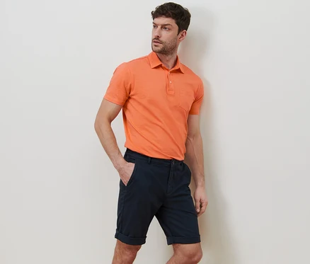 Un homme pose en short chino bleu marine et polo orange.