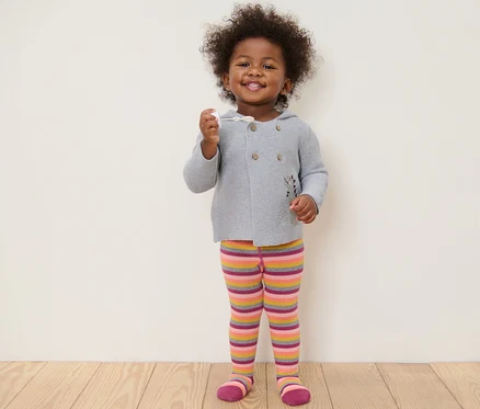 Un enfant souriant debout tenant un jouet. Il porte un pull gris et des collants roses rayés.