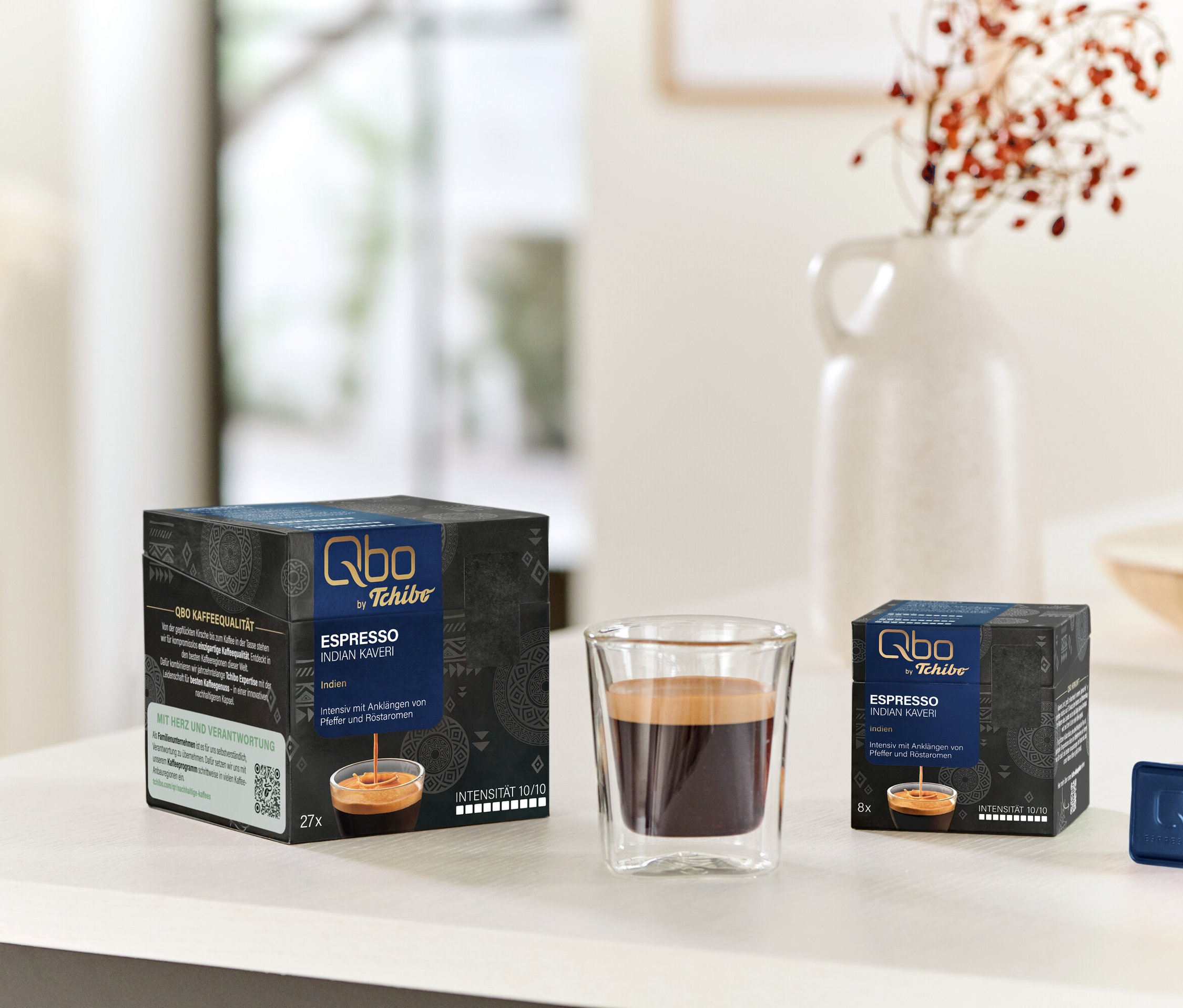 Sur une table, une tasse d'espresso et des paquets de Qbo Espresso INDIAN KAVERI avec 27 et 8 capsules.