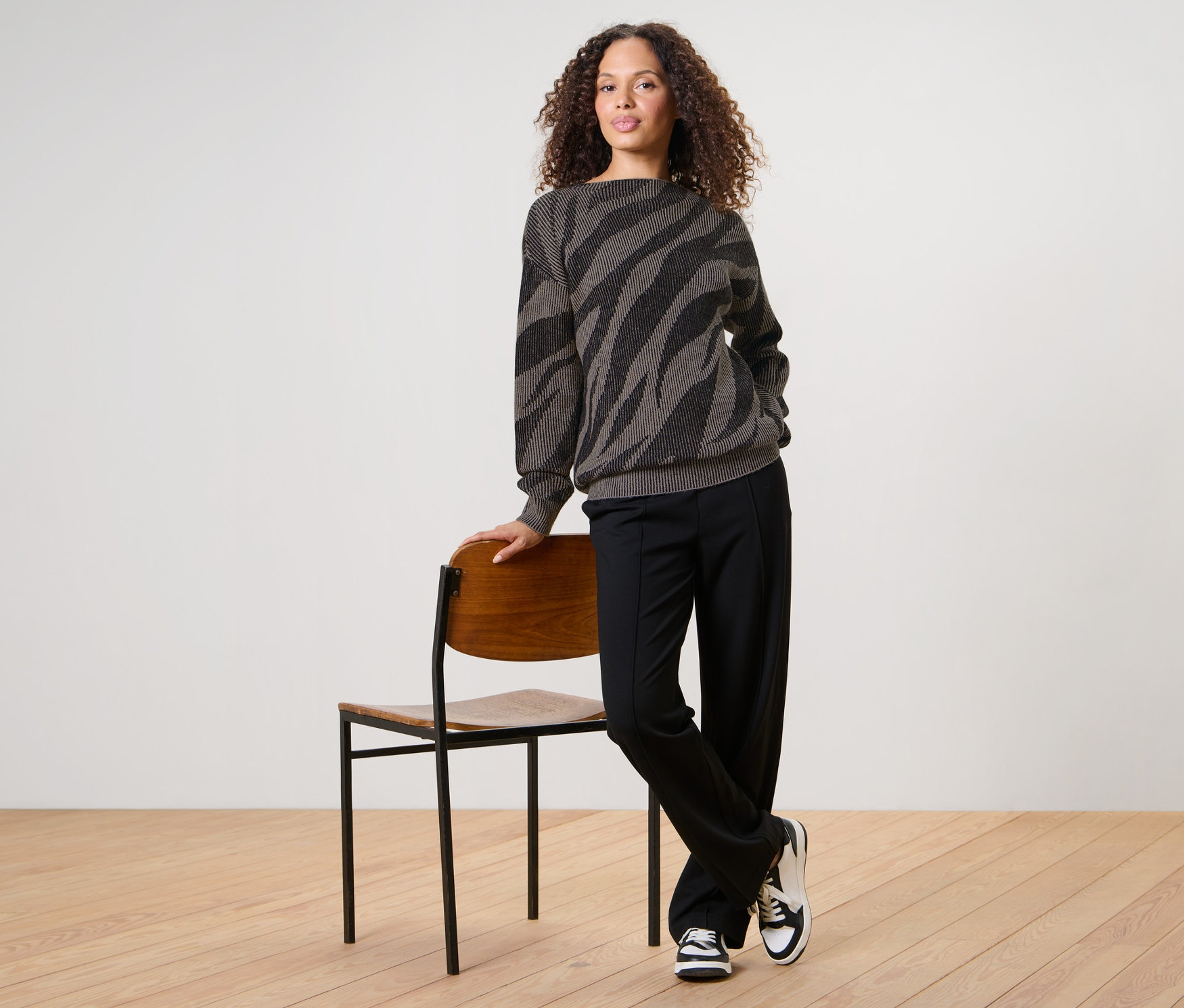 Une femme pose, appuyée contre une chaise, portant un pull en maille jacquard.