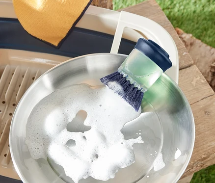 Une poêle en acier inoxydable est lavée avec une brosse avec distributeur. À côté se trouvent un égouttoir et un chiffon.