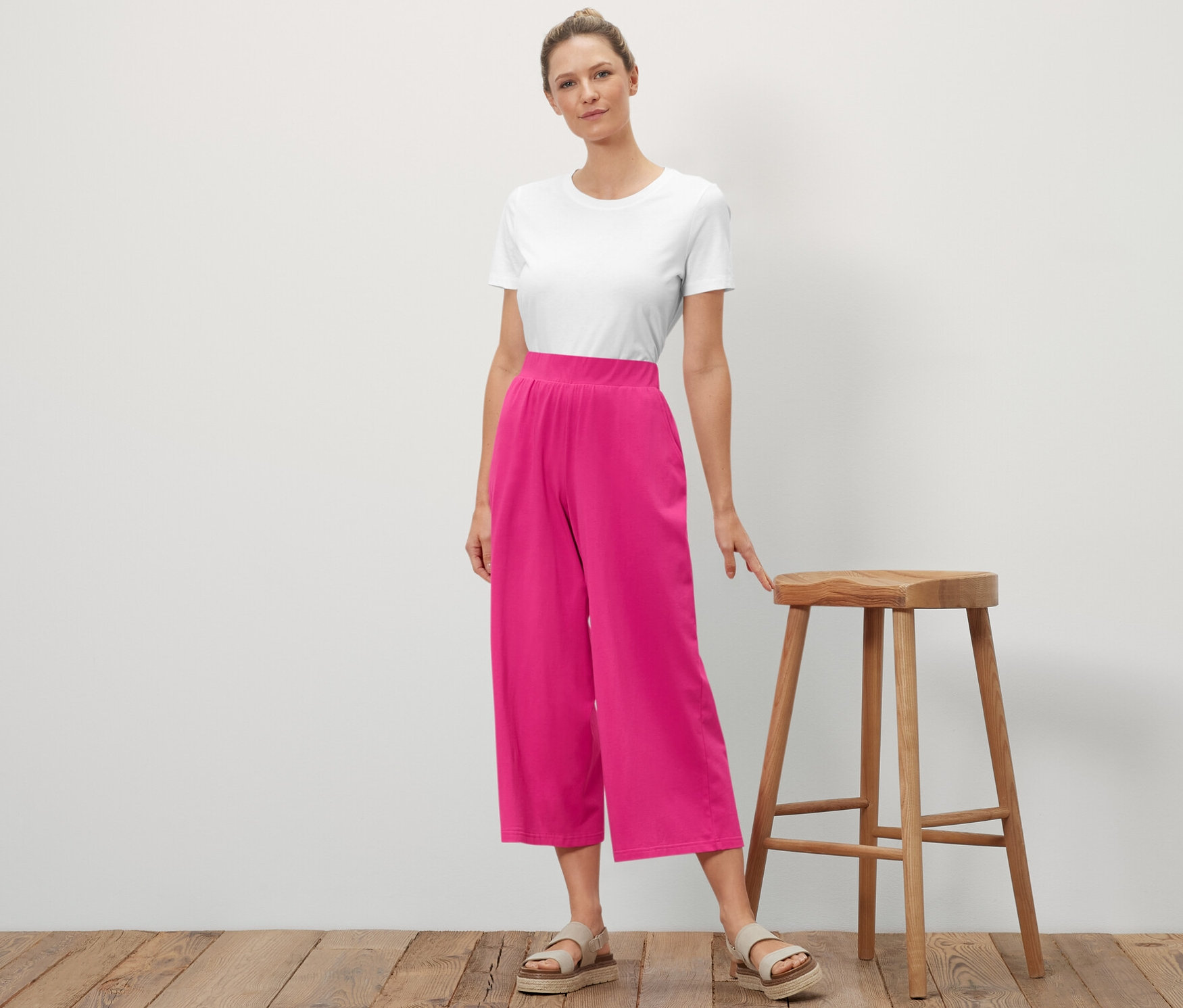 Une femme se tient à côté d'un tabouret en bois et pose. Elle porte un t-shirt blanc et un pantalon jersey rose.