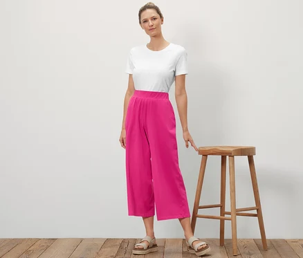 Une femme se tient à côté d'un tabouret en bois et pose. Elle porte un t-shirt blanc et un pantalon jersey rose.