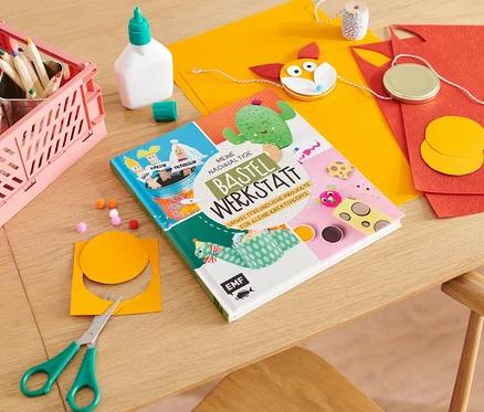 Sur une table sont posés le livre « Meine nachhaltige Bastelwerkstatt » (en allemand), des ciseaux, des papiers colorés, de la colle et des crayons de couleur.
