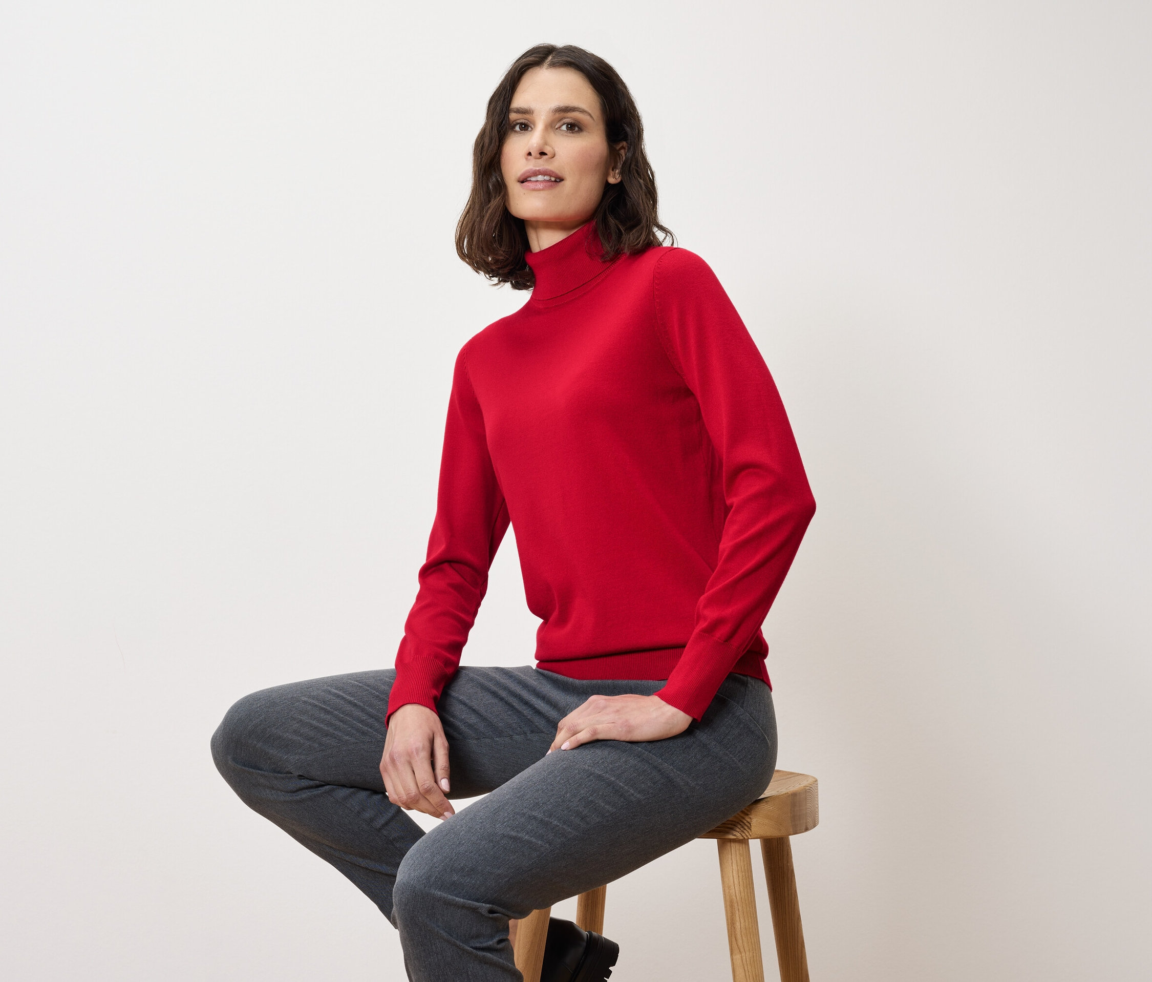 Une femme est assise sur un tabouret, portant un pull à col roulé rouge et un pantalon gris.