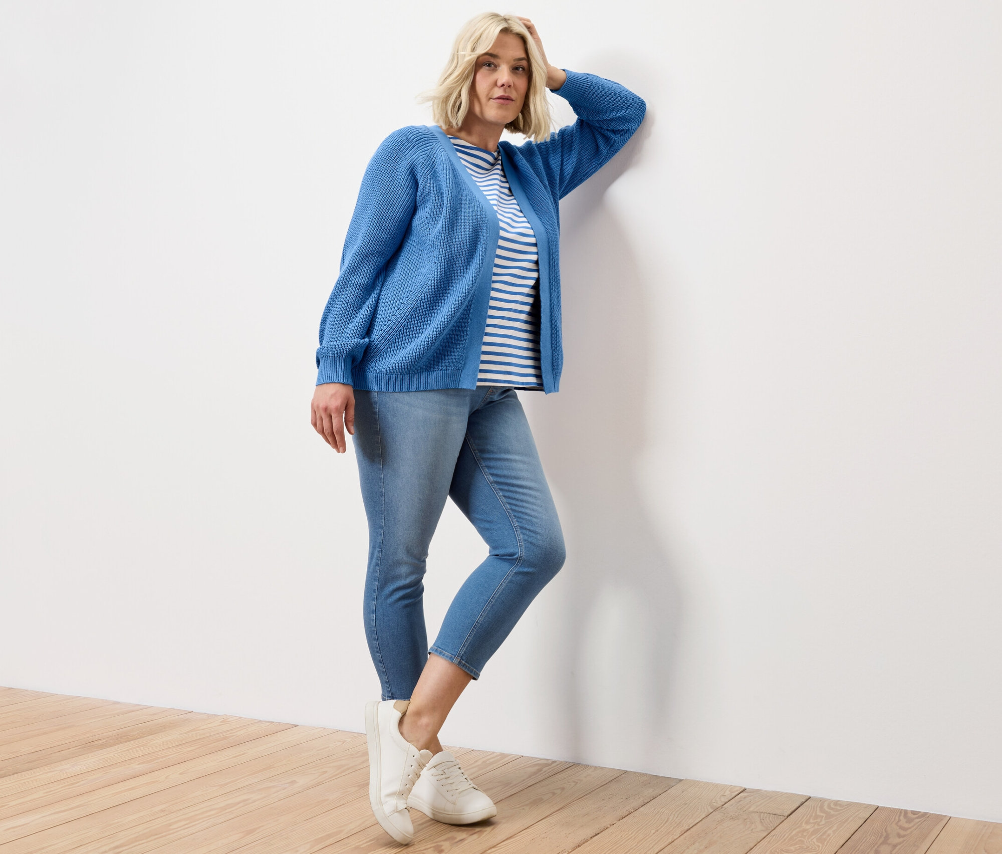 Une femme pose, appuyée contre un mur, portant un t-shirt bleu à rayures à demi-manches, un tregging 7/8 – Fit « Nelly » et un cardigan ouvert.