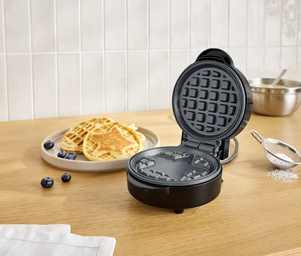 Gaufrier noir ouvert à côté d'une assiette de gaufres et de myrtilles sur une table en bois.