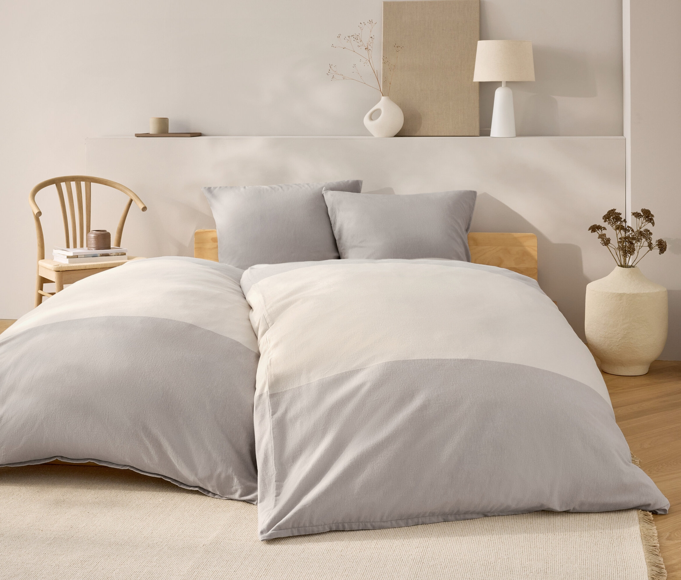 Chambre avec un lit avec des draps gris et blancs, deux oreillers gris et une tête de lit en bois. À l'arrière-plan, une chaise en bois et des décorations.