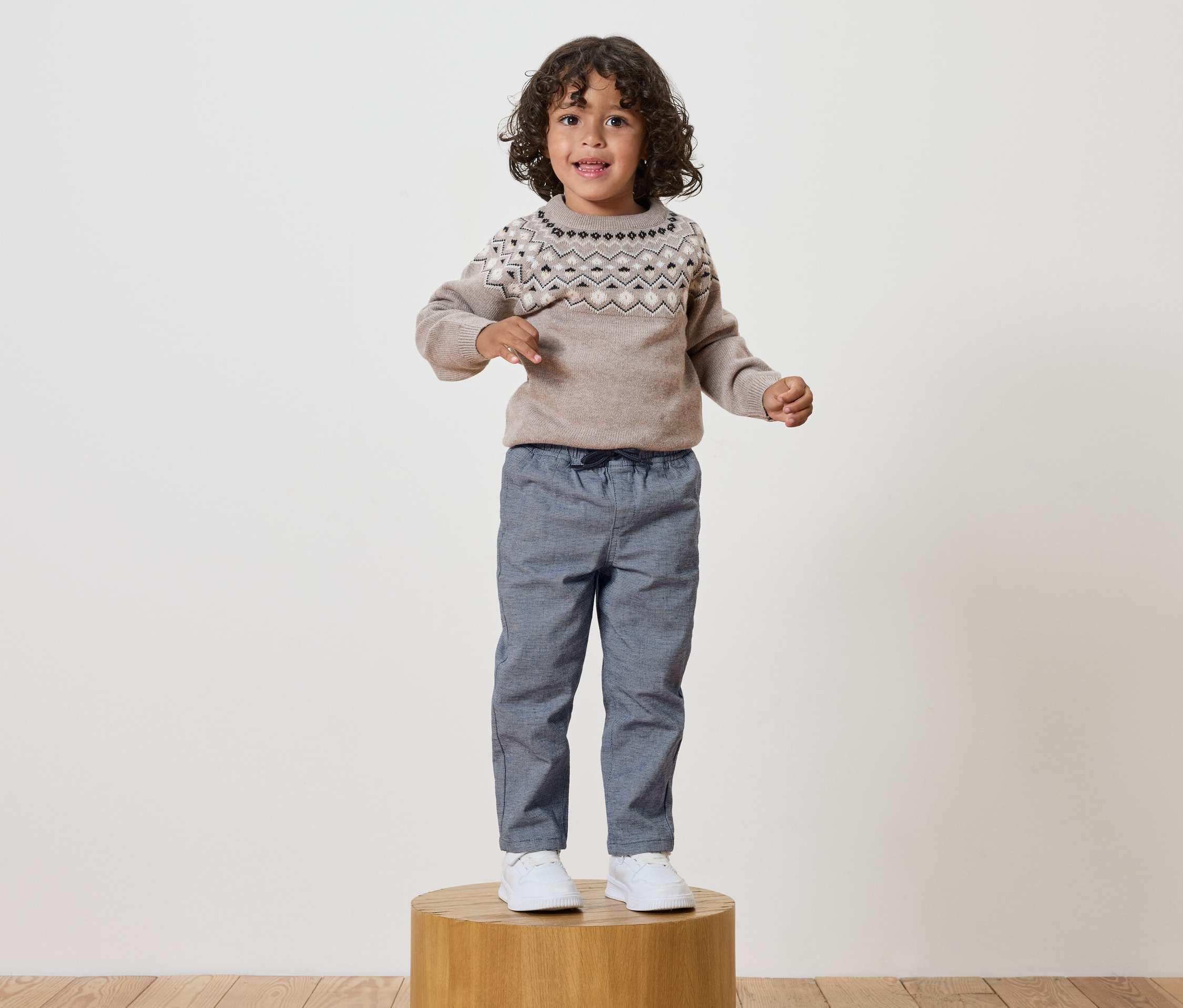 Un garçon aux cheveux bouclés se tient sur un socle en bois, portant un pull à motifs et un pantalon bleu.