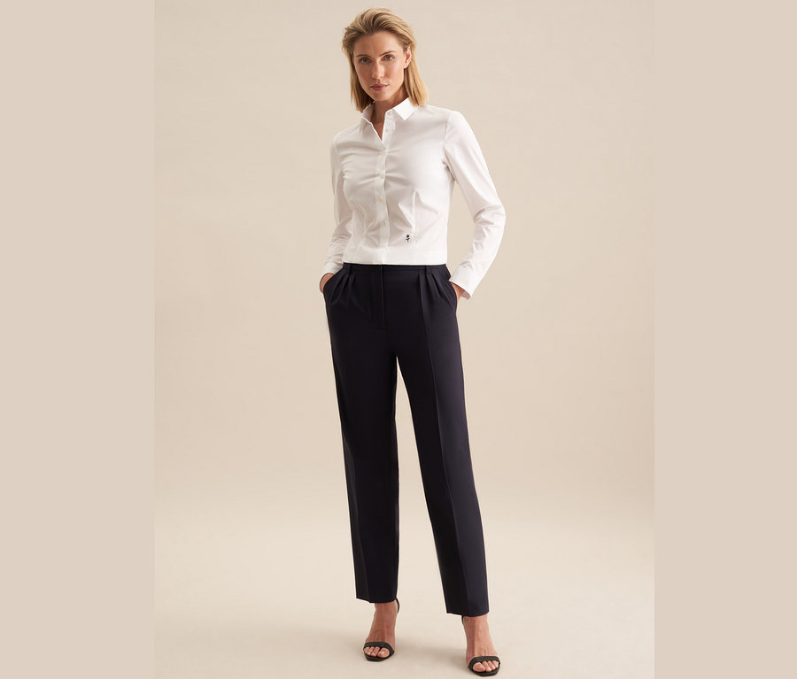 Une femme se tient debout, les mains dans les poches de son pantalon. Elle porte un chemisier Seidensticker Slimfit blanc, un pantalon bleu marine et des sandales noires.
