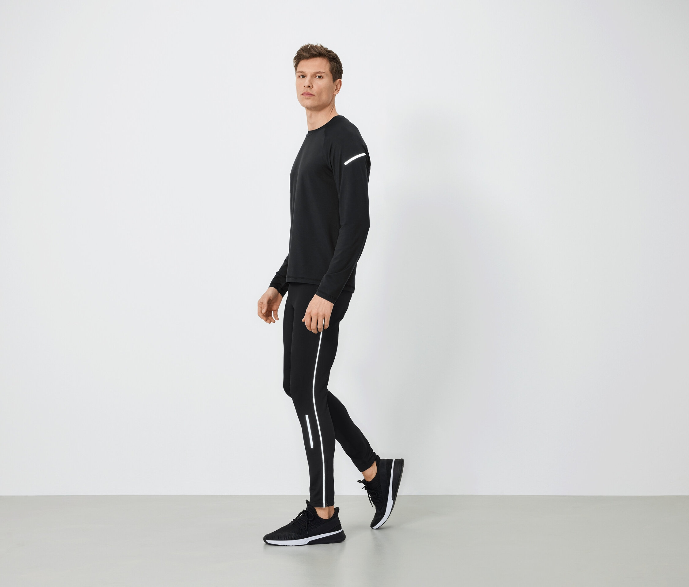 Un homme en tenue de sport noire avec des détails réfléchissants marche sur un sol gris.