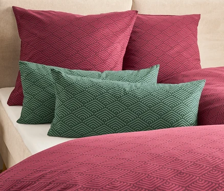 Sur un lit, deux oreillers bordeaux et deux oreillers verts avec un motif géométrique.