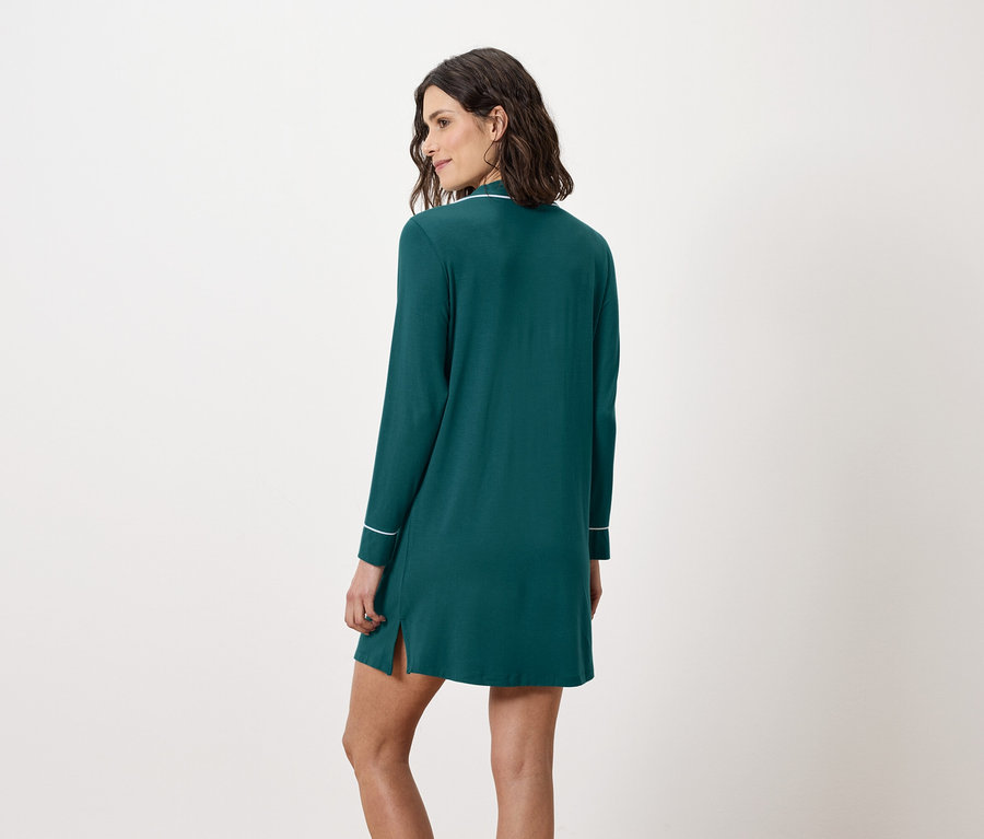 Une femme se tient dos à la caméra et porte une chemise de nuit vert émeraude à manches longues avec des bordures blanches.