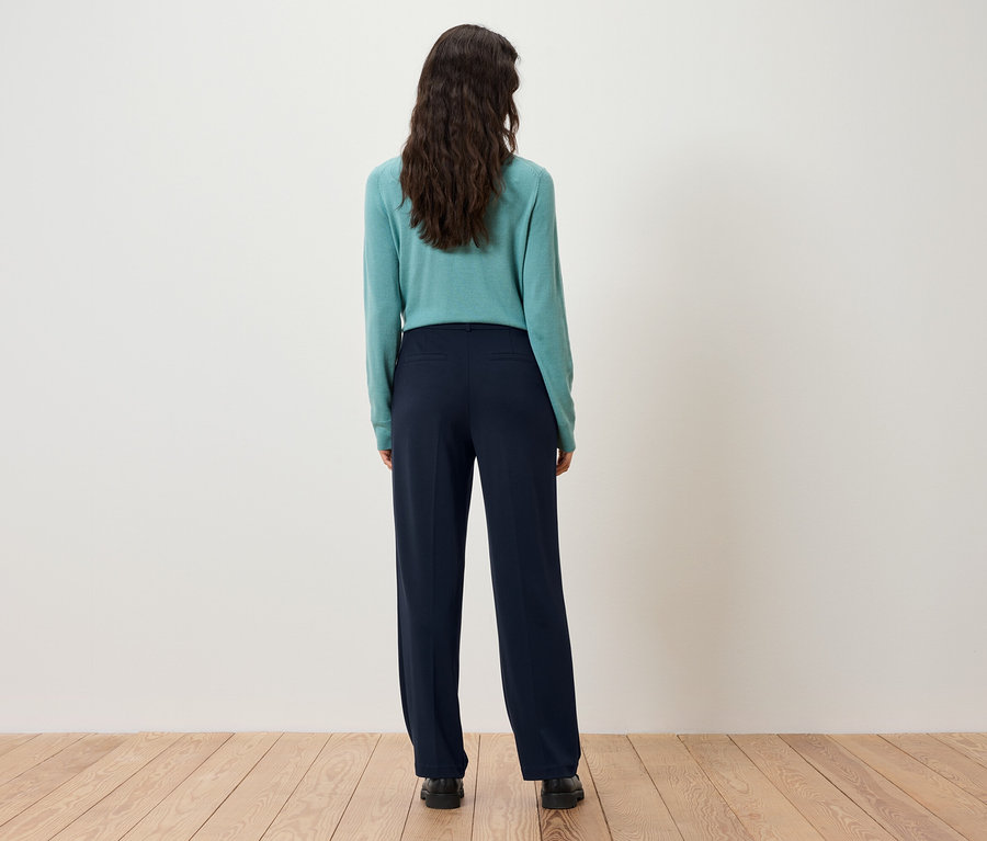 Vue arrière d'une femme aux cheveux châtain foncé portant un pull turquoise et un pantalon bleu marine.