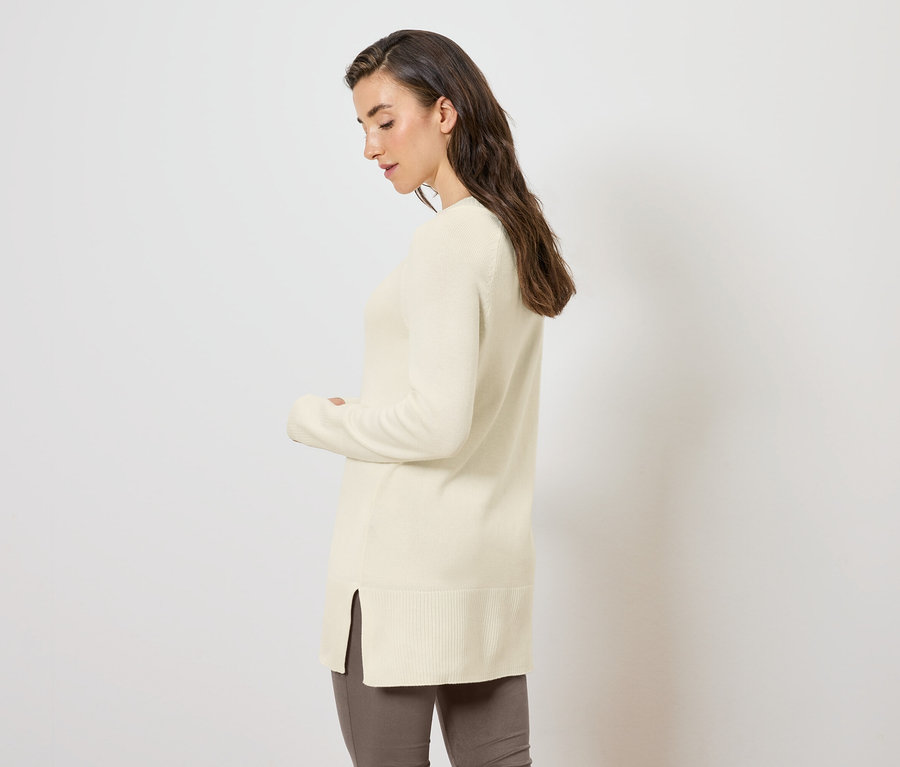 Femme posant de côté dans un long pull en maille crème.