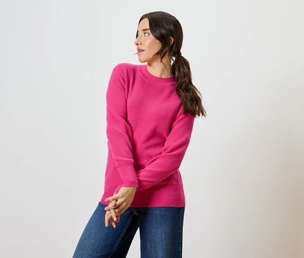 Une femme aux cheveux bruns et aux yeux bleus pose dans un pull rose et un jean bleu.
