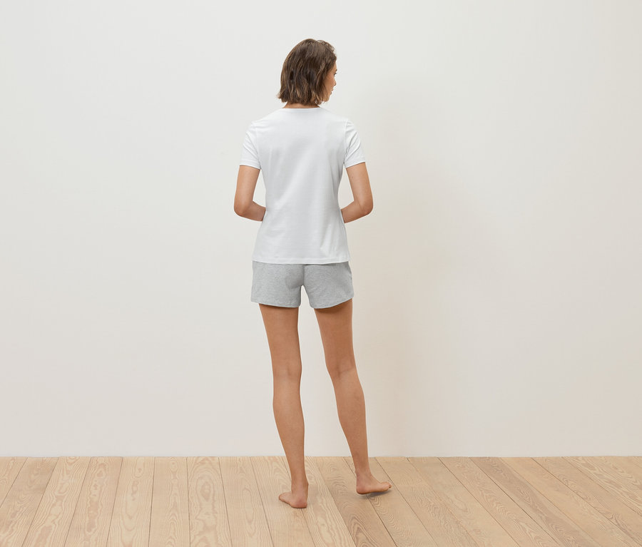 Une femme vue de dos avec des cheveux bruns courts portant un t-shirt blanc et un short gris. Elle se tient sur un plancher en bois devant un mur blanc.