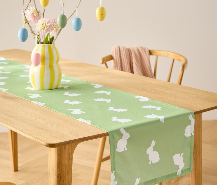 Table à manger avec un chemin de table vert clair à motifs de lapins blancs, un vase avec des fleurs et une décoration d'œufs de Pâques.