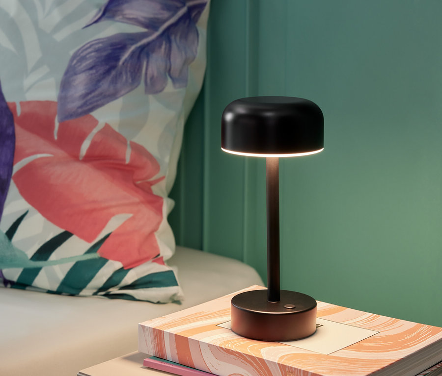 Une lampe de table à LED noire sans fil posée sur un livre à côté d'un coussin.