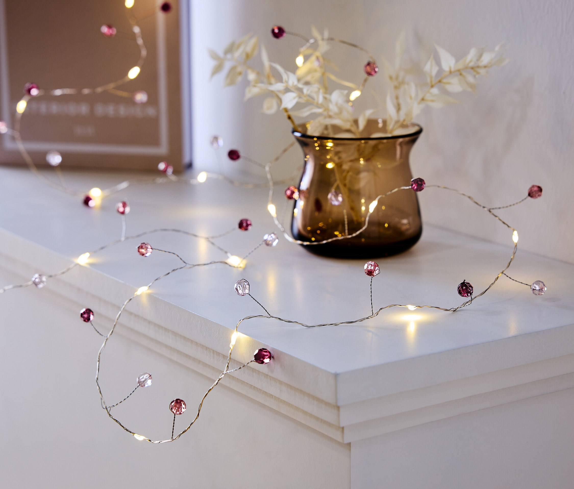 Une guirlande lumineuse décorative avec des perles violettes et transparentes est posée sur une étagère blanche à côté d'un vase avec des plantes et un livre.