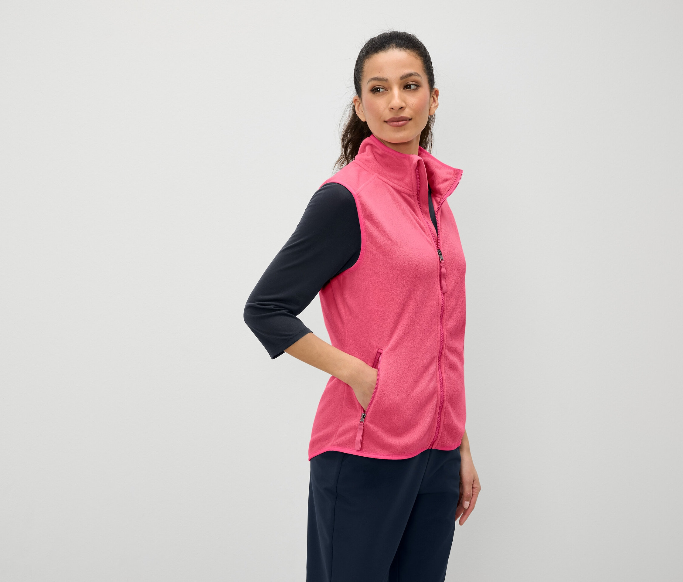 Une femme aux cheveux bruns a la main dans la poche d'un gilet rose. Elle porte un t-shirt bleu marine à manches trois-quarts et un pantalon bleu marine.