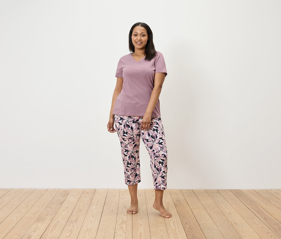 Une femme se tient debout et porte un t-shirt violet et un pantalon à fleurs.