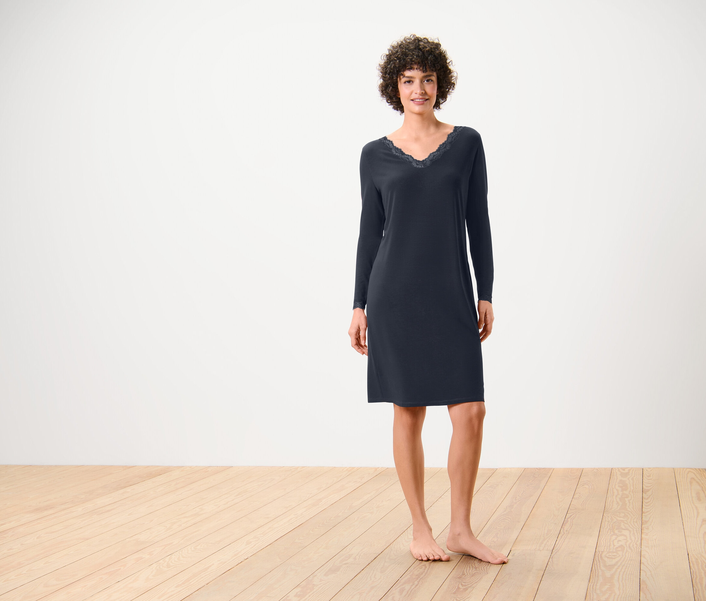 Une femme se tient pieds nus sur un plancher en bois, elle porte une chemise de nuit bleu marine à manches longues avec de la dentelle à l'encolure.