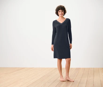 Une femme se tient pieds nus sur un plancher en bois, elle porte une chemise de nuit bleu marine à manches longues avec de la dentelle à l'encolure.