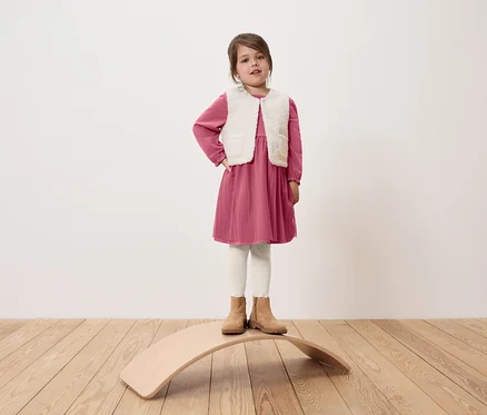 Une fille se tient sur une planche d'équilibre en bois, elle porte une robe rose, un gilet blanc et des bottes beiges.