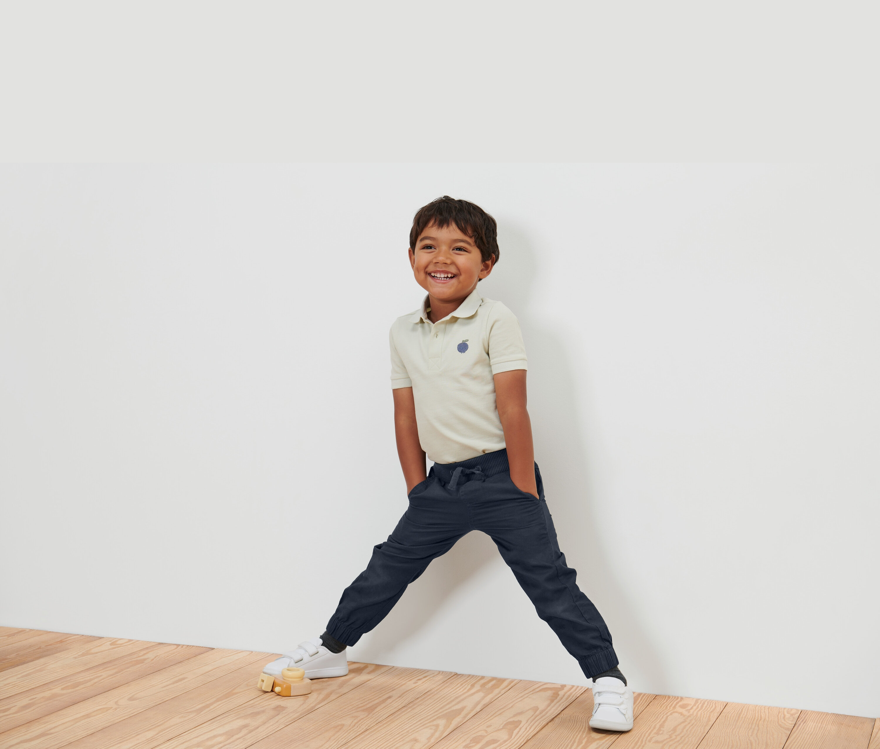 Un garçon souriant portant un polo beige, un pantalon bleu marine et des chaussures blanches, se tient contre un mur blanc.