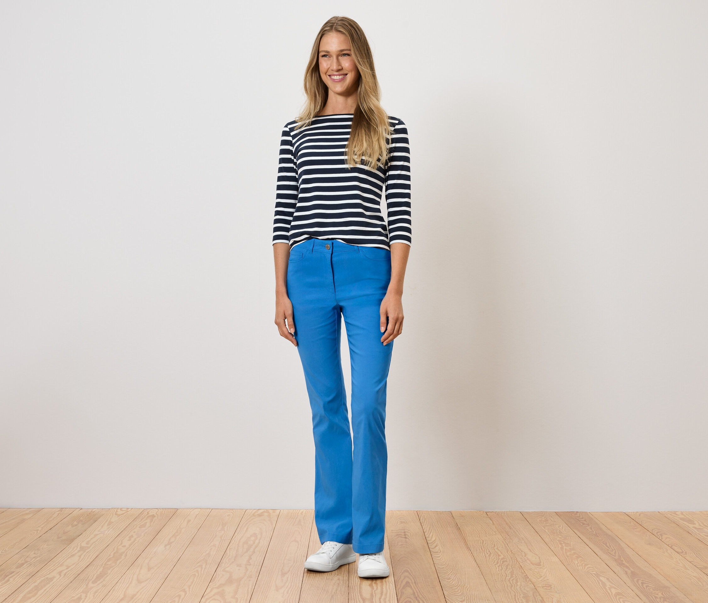 Une femme debout porte un pantalon bleu, un t-shirt rayé et des chaussures blanches.