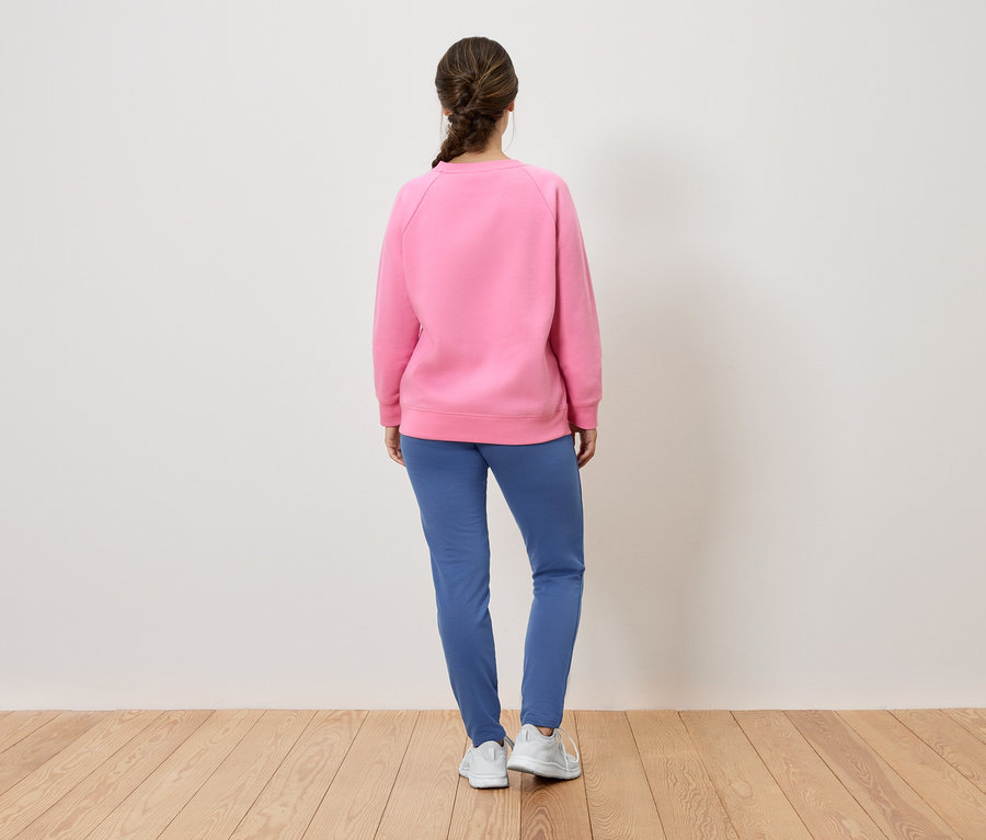 Une femme, dos à la caméra, porte un sweat-shirt rose, un pantalon bleu et des chaussures blanches.